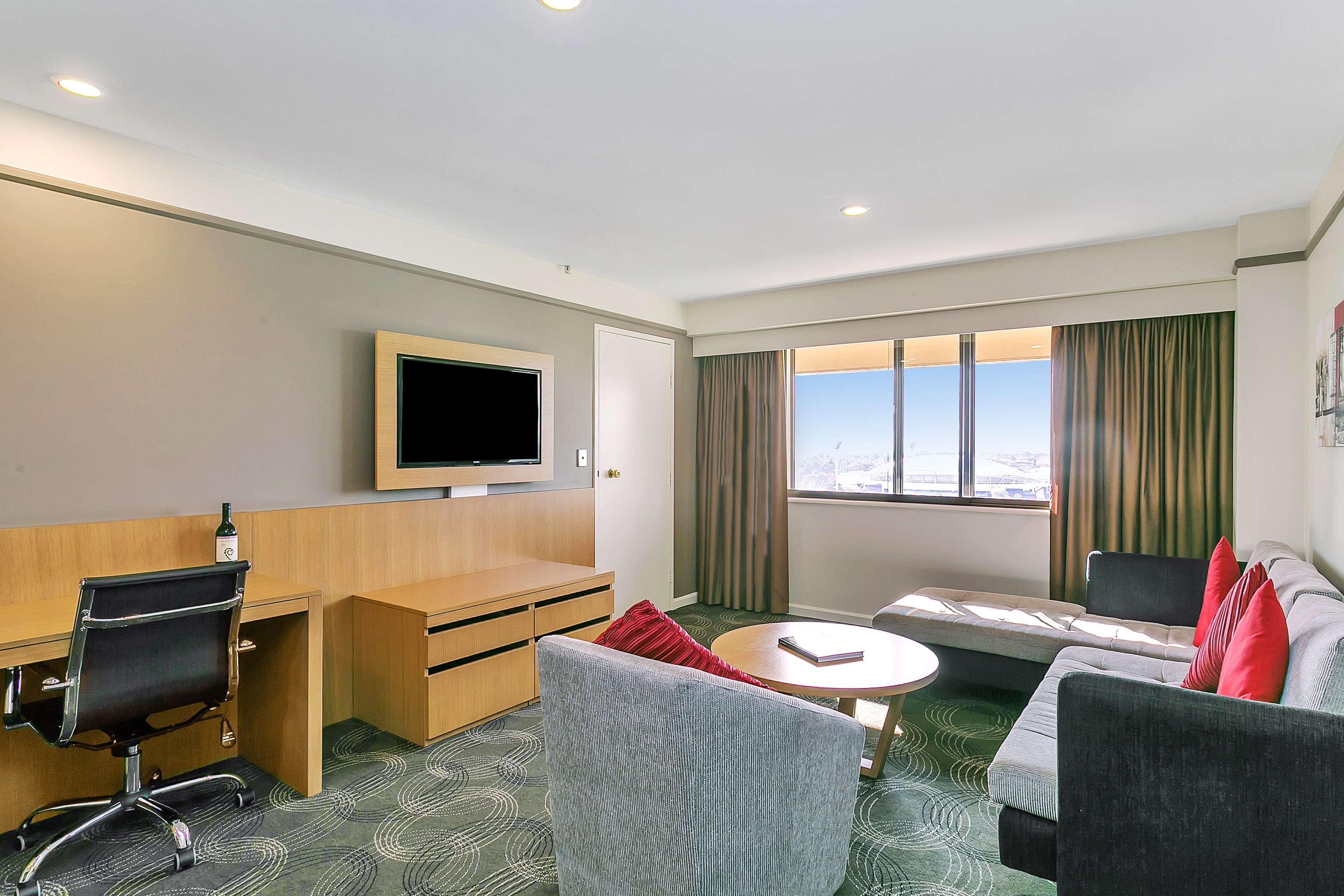Stamford Plaza Adelaide Hotel Ngoại thất bức ảnh The photo shows a modern hotel room or suite that features a cozy seating area. There is a comfortable sofa with red accent pillows, a round coffee table in the center, and a sleek desk with an office chair. On the wall, there is a flat-screen TV mou